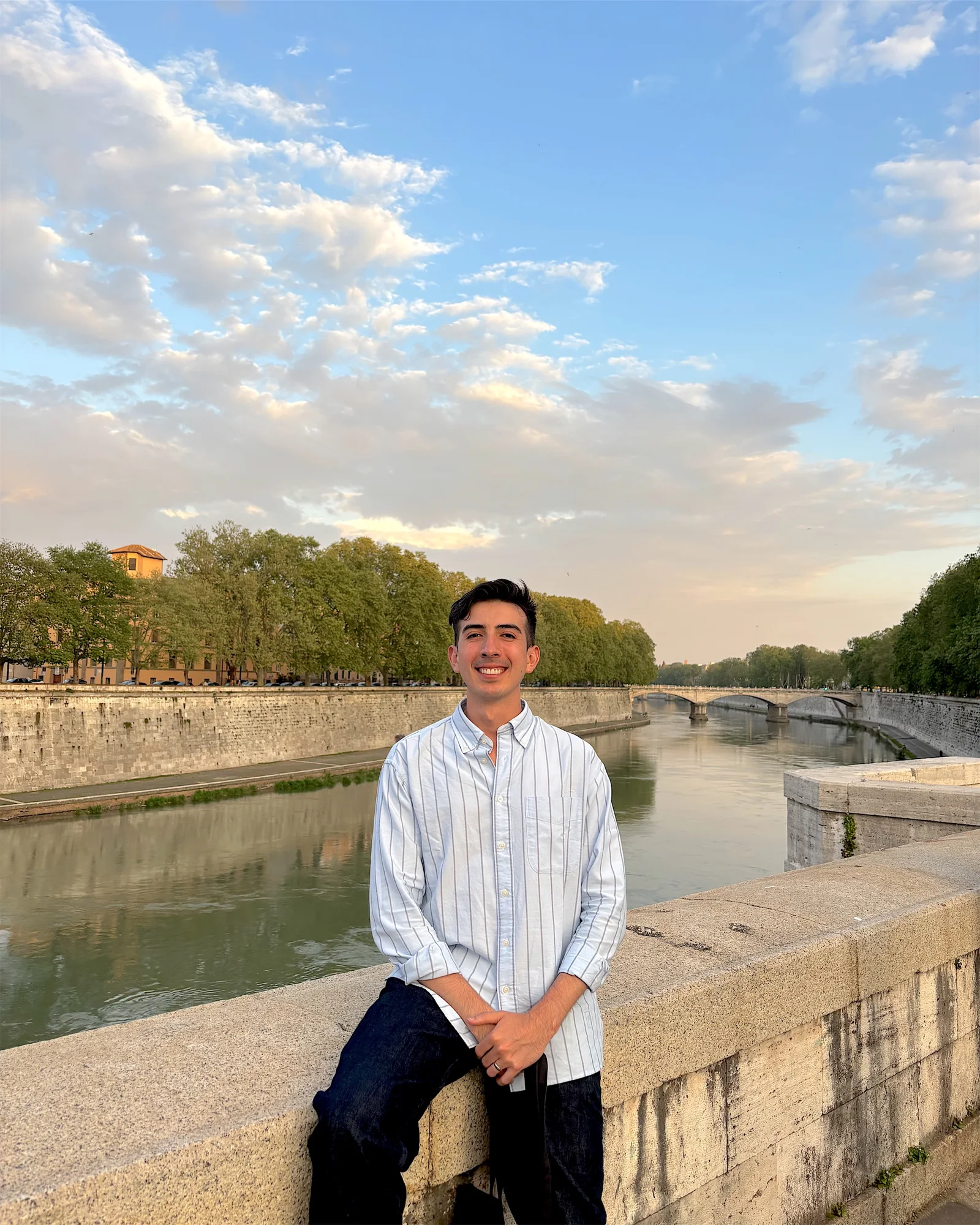 David Zambrano smiling, seated on a stone wall beside the Tiber in Rome at dusk, wearing a light striped shirt.